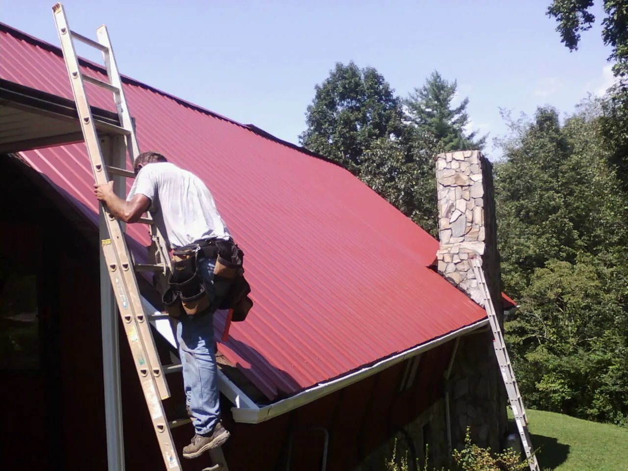 Roofer on a ladder beside a red metal roof and stone chimney — Soffit & Fascia Repair by Roofing Brothers in Crystal Springs